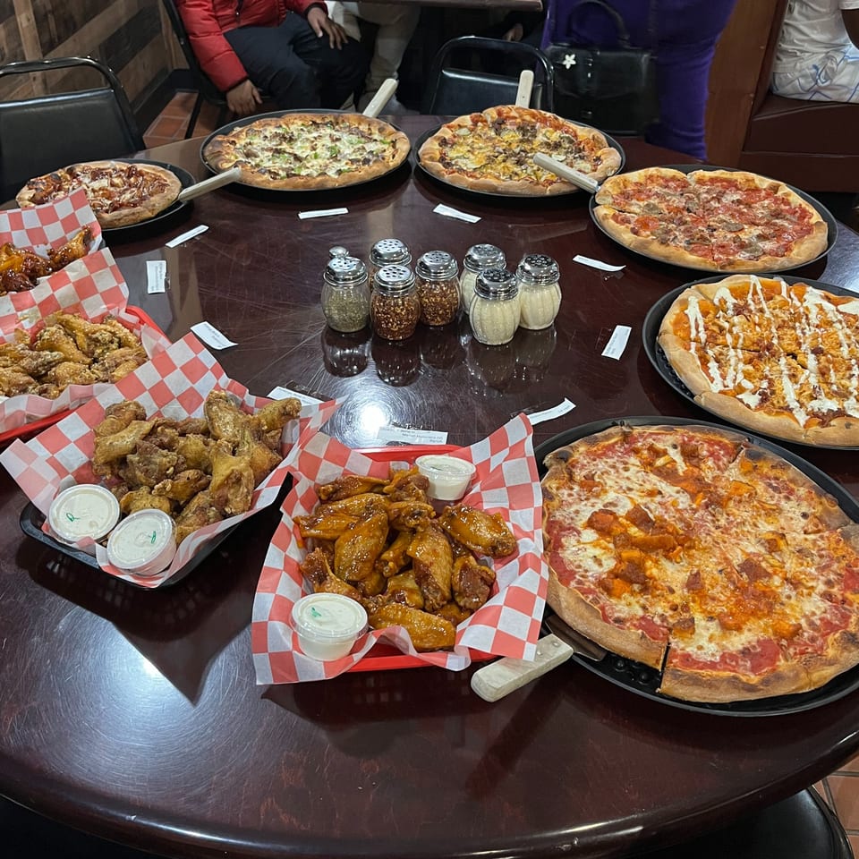 A round table laden with six Bodacious Pizza & Grill pizzas and three baskets of chicken wings.
