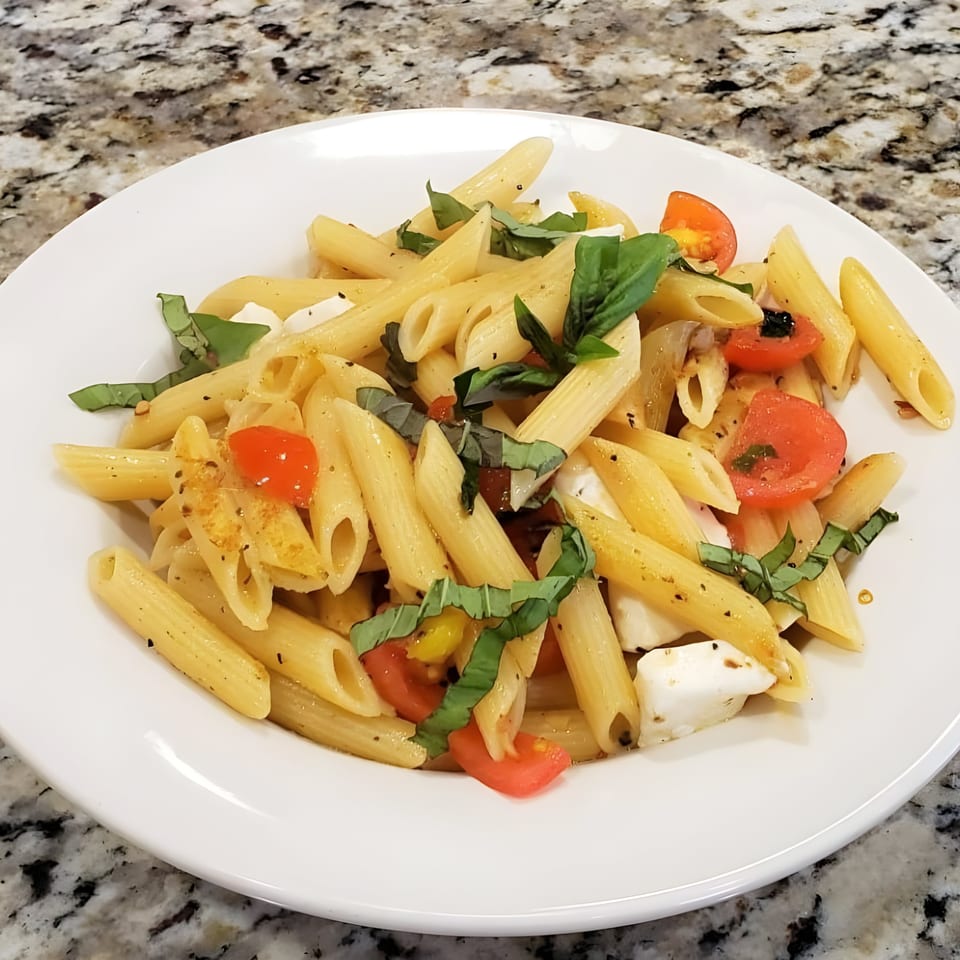 Penne pasta salad with cherry tomatoes, fresh mozzarella, and basil on a white plate.